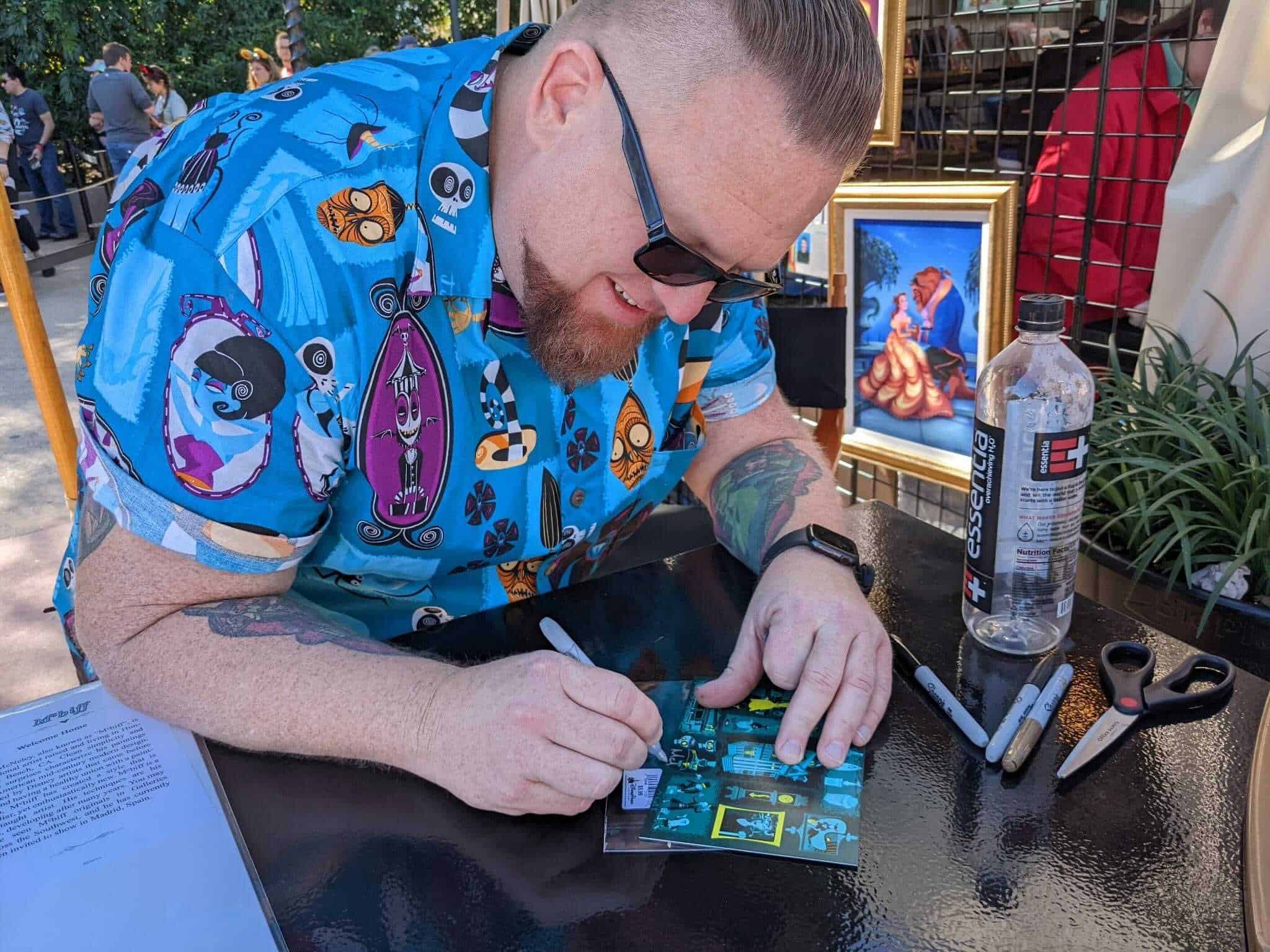 Artist McBiff signs a Haunted Mansion tiki themed piece of art on a postcard at Epcot Art festival