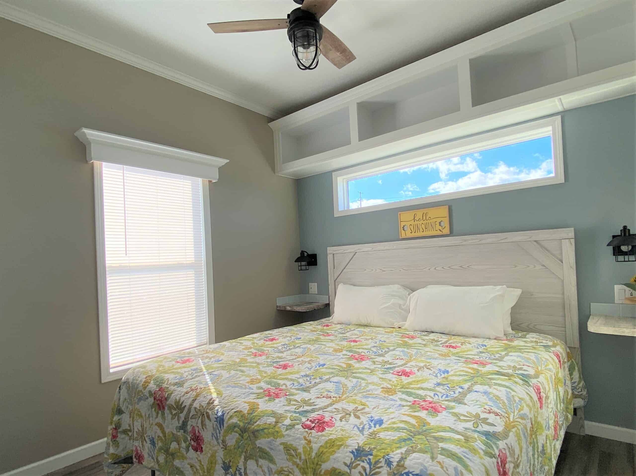 Cabana Club Cottage Bedroom with King Bed has gray and blue soft tone painted walls, a floral comforter blanket and white pillows on the bed with a white wood headbord and a long window show blue skies above it, and a yellow sign that reads "hello sunshine" above the headboard