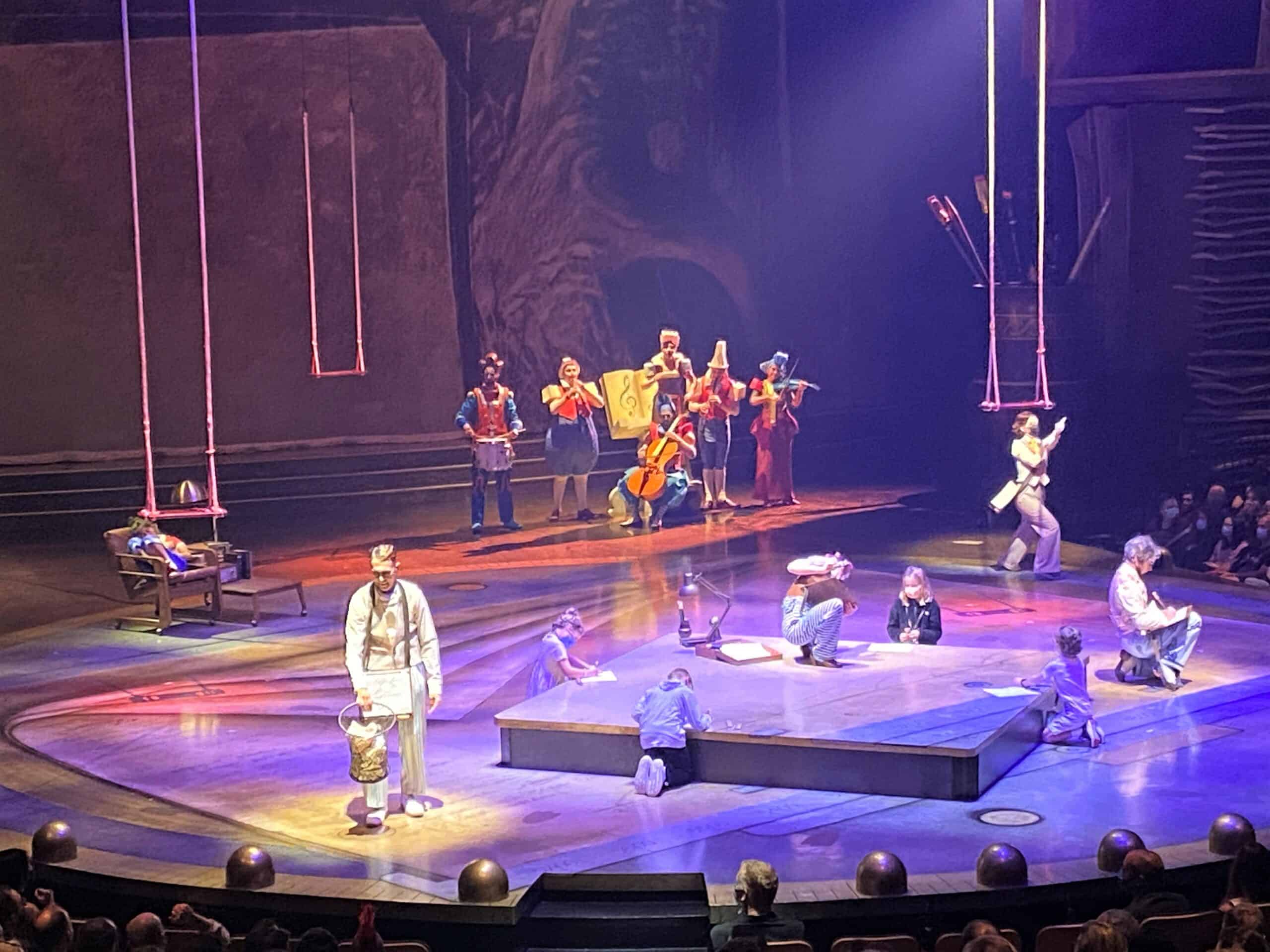 Cirque du Soleil Disney Springs - Preshow involves multiple performers on stage as well as audience participating, including from kids in the audience. The stage is illuminated in rich metallic and purple tones. Multiple performers are on stage and a small group of children are sitting around a square on the stage, drawing pictures. 