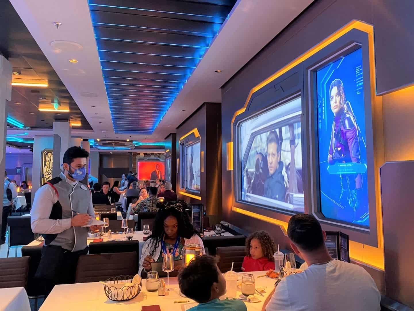 Dining Room of Worlds of Marvel Restaurant on Disney Wish with Marvel characters on screens around the walls of the restaurant 