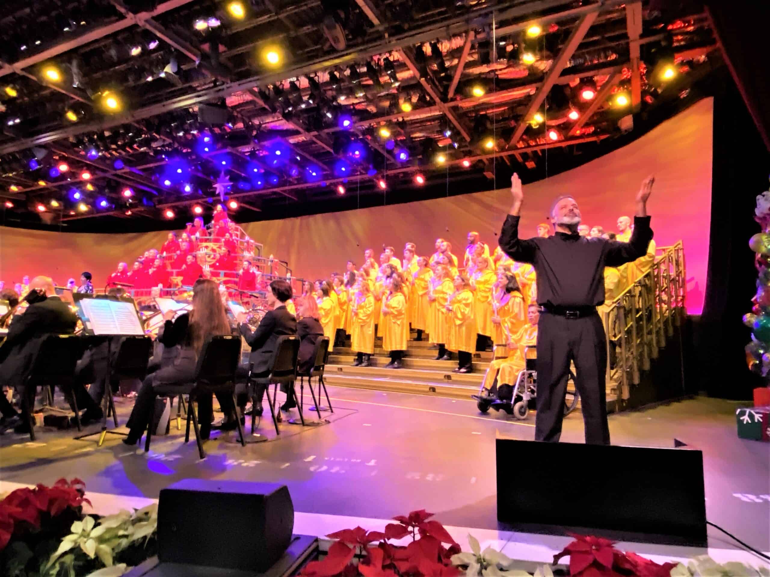 EPCOT Candelight Processional Returns for 2021- in this photo of the stage a sign language interpreter is near the front of the stage, dressed in all black with his arms raised as he inteprets the music being performed by an orchestra to his right, dressed in all black, a choir dressed in all red staged elevated above the orchestra, and a larger choir dressed in golden gowns staged on bleachers behind the interpreter. 
