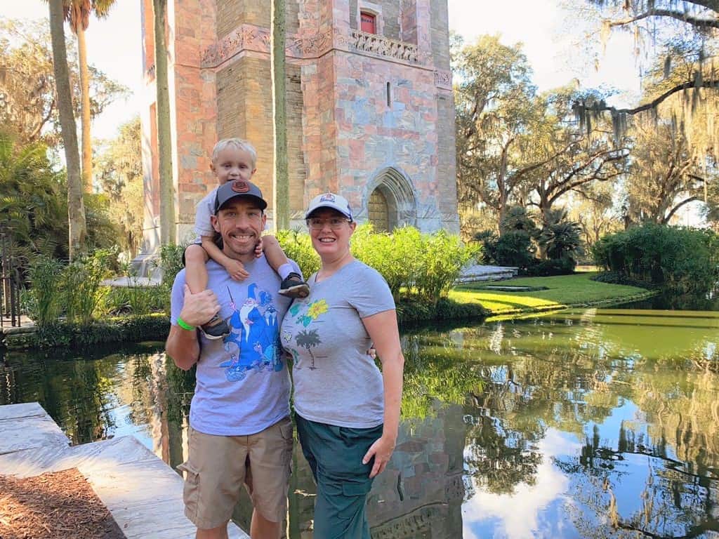 Family Time at Bok Tower Gardens - a family of three pose facing the camera, each wearing a t-shirt and shorts, as the Bok Tower stands behind them and its reflecting pool shows the reflection of green trees and blue skies