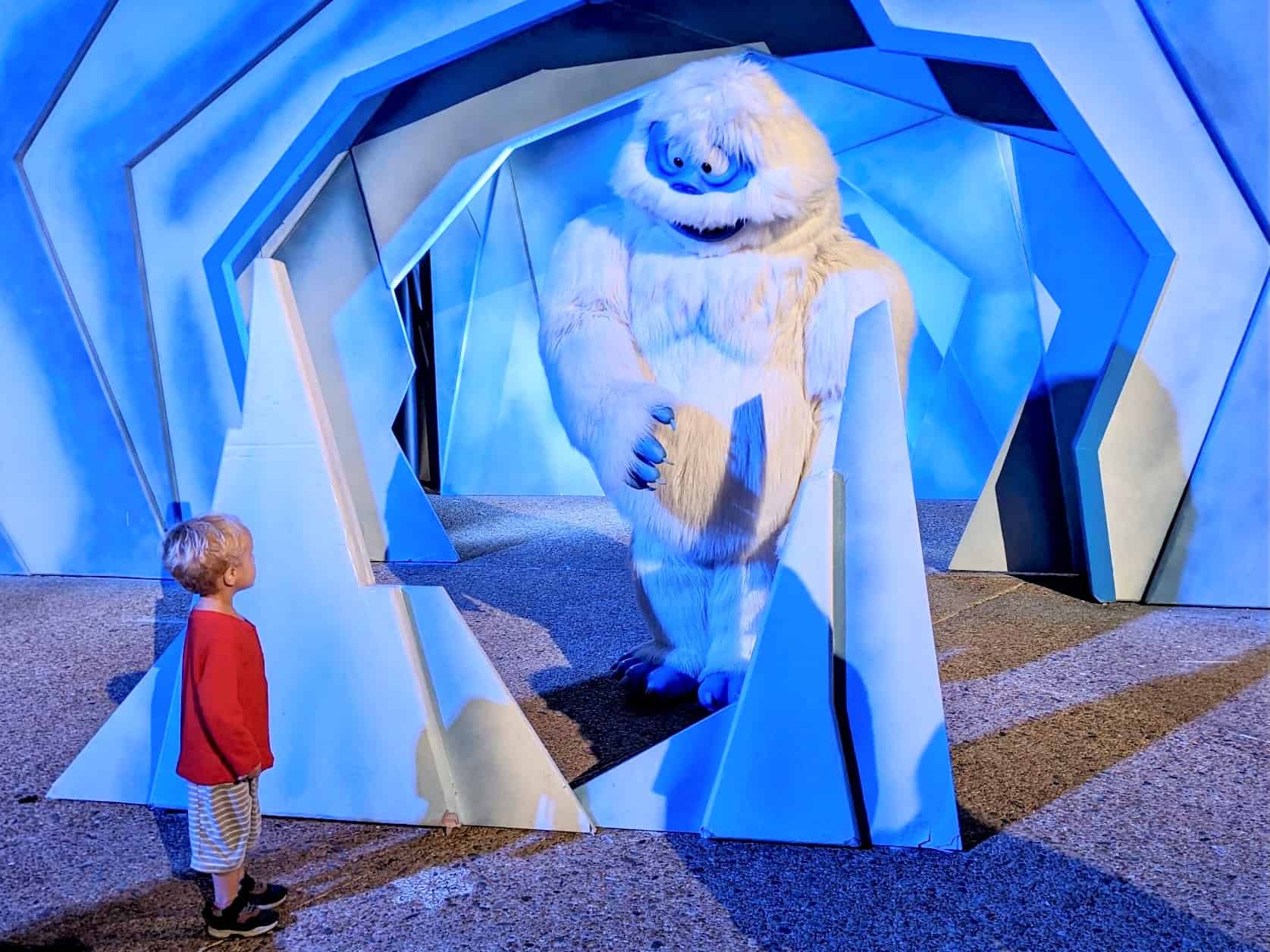 Meeting Bumble from Rudolph the Red Nosed Reindeer at SeaWorld Orlando. A large costumed character has white fur and long limbs with a blue face and hands, looks down at a three year old boy facing him, wearing a red long sleeve shirt and grey shorts. There is a white and blue background to resemble an ice cave. 