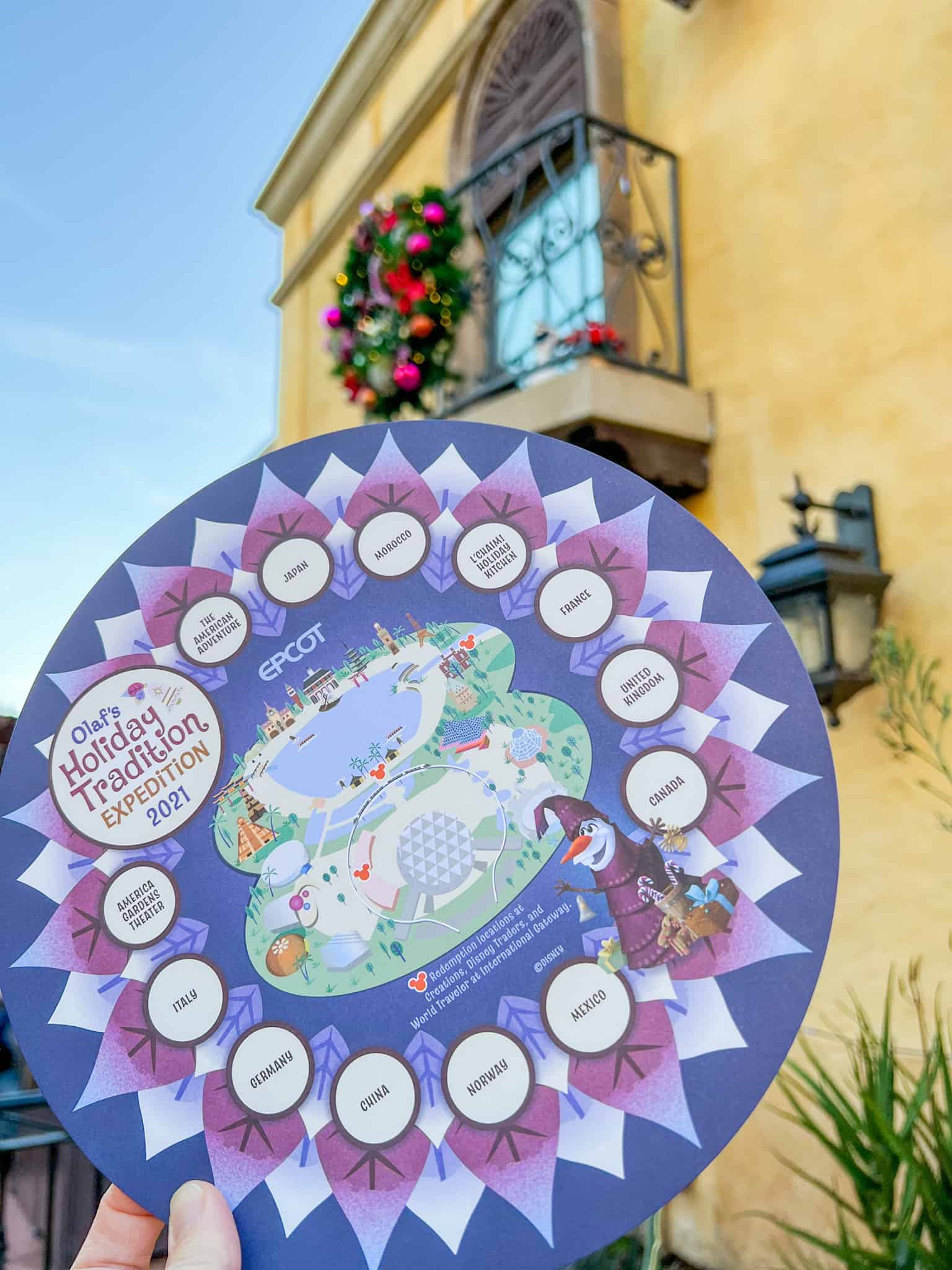 A circular map of the Olaf's Holiday Traditions Expedition - EPCOT Festival of the Holidays is held in front of the camera with a tan hued building of the Mexico pavilion in the background. On the balcony of this building is a small Olaf character statue and a festive wreath.