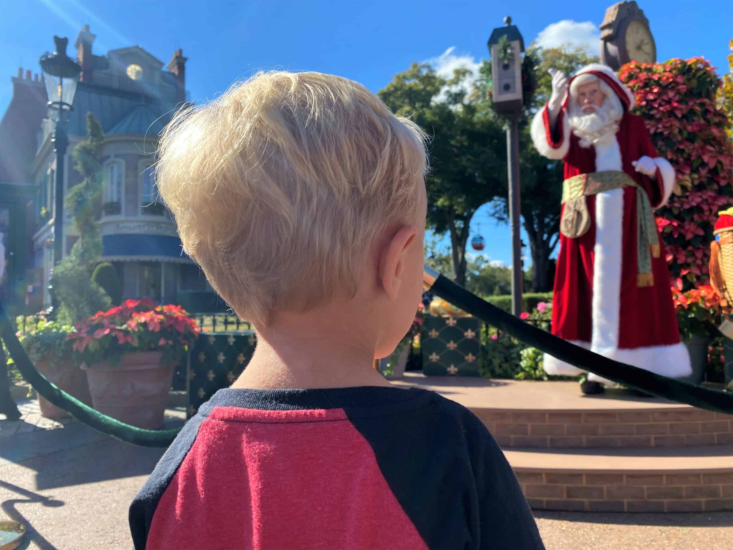 Pere Noel Holiday Storyteller at EPCOT France Pavilion stands on a small stage wearing a winter coat with deep red colored fur and white trim. In the foreground a three year old boy with blonde hair and a red long sleeve shirt watches the show.