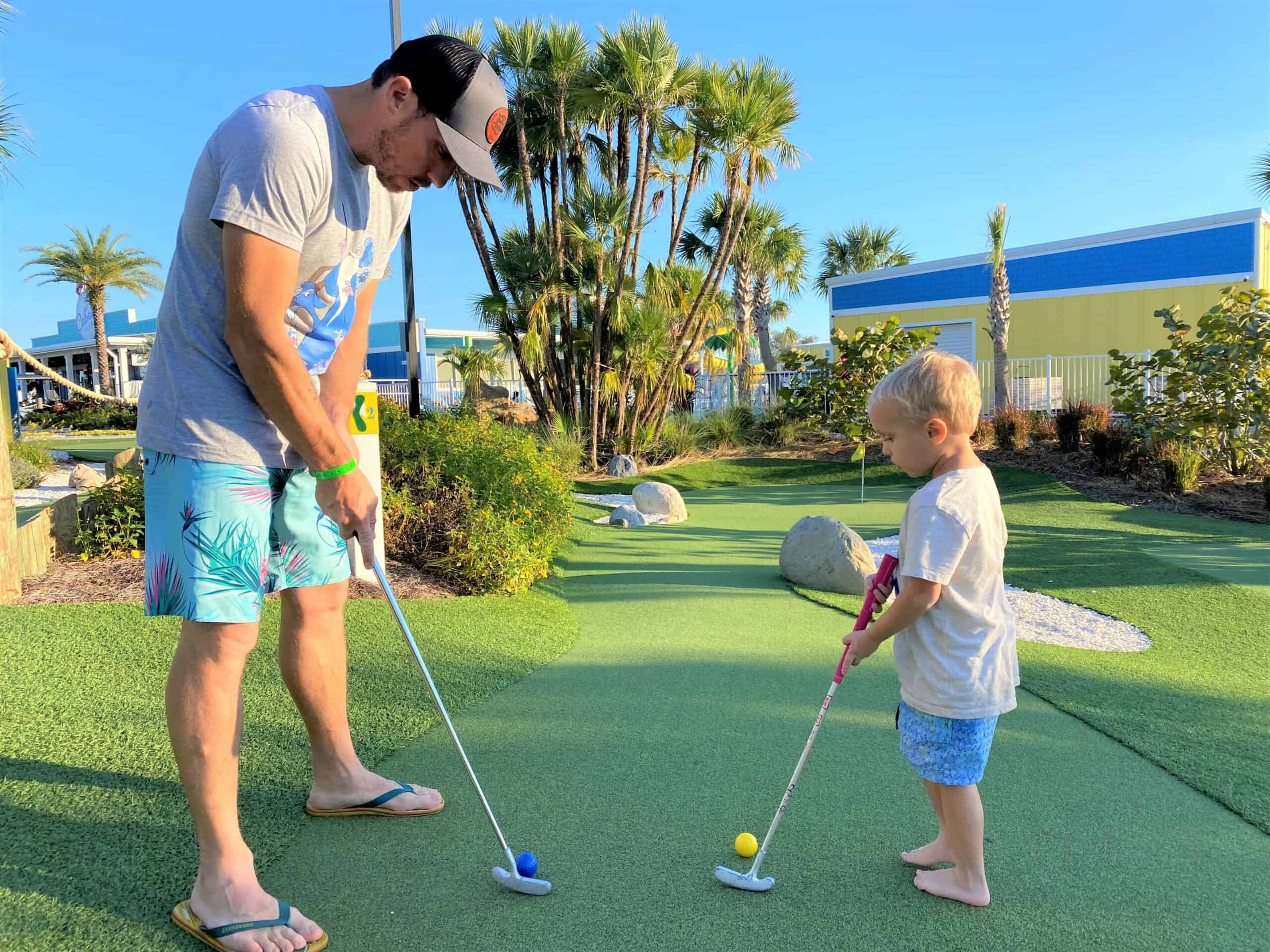 A father in gray t-shirt, baseball hat, and wearing tropical print shorts and flip flops holds a putting tee in his hands as his four year old son faces him and also has a putting tee in his hands, as they are both about to put a golf ball down the green at Putting Course at Cabana Club Resort