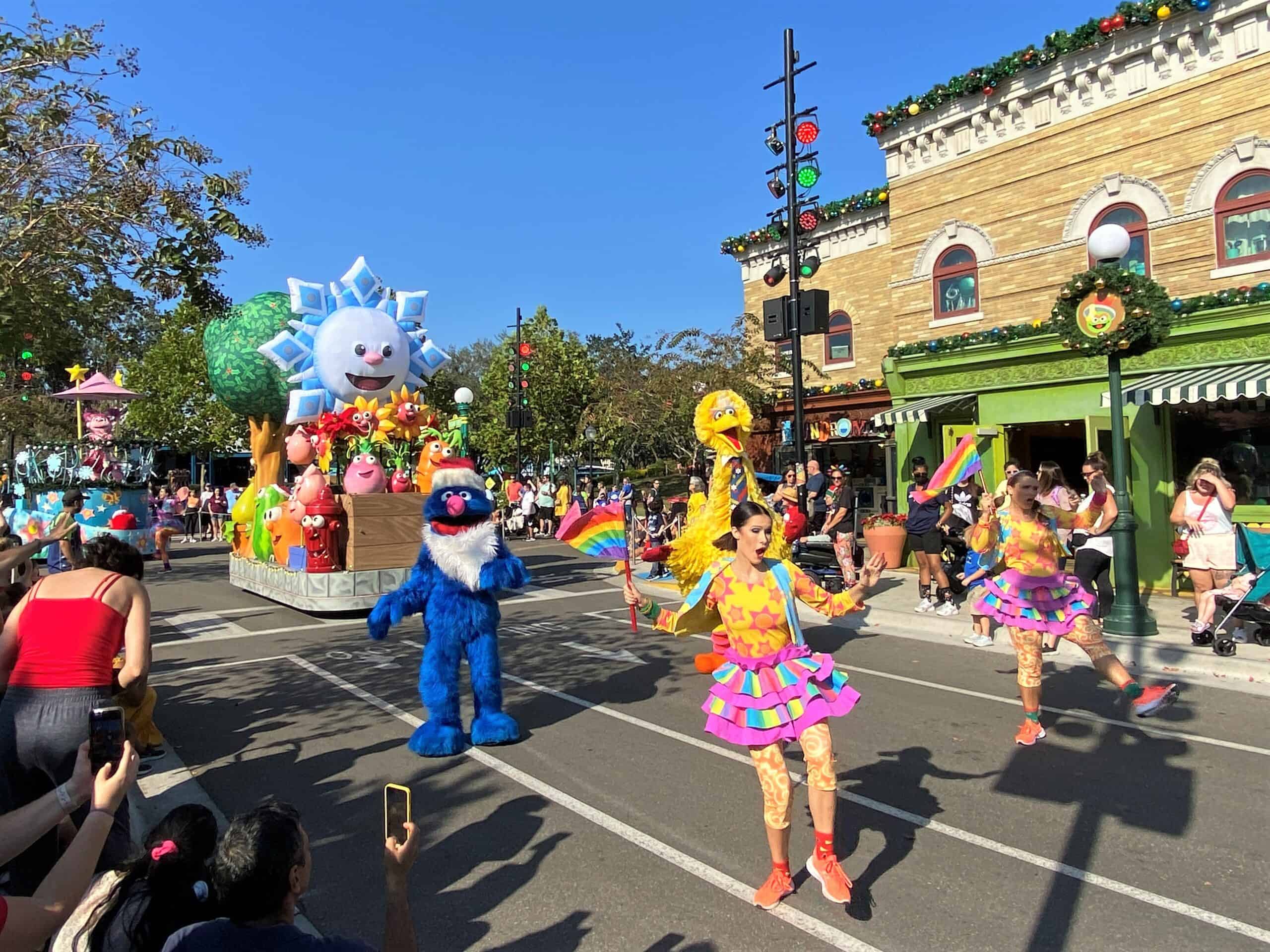 This photo is taken during the day inside Sesame Street Land at SeaWorld Orlando. Two female performers lead the parade dancing and wearing bright colored costumes of yellow and pink. Big Bird and Grover characters are behind them, and a large parade float with bright flowers and a large inflated snowflake are behind them.Sesame Street Christmas Parade
