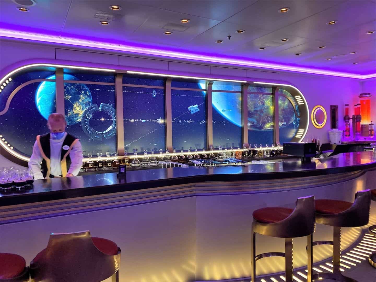 Star Wars Hyperspace Lounge for Adults on Disney Wish - a bartender stands behind an elegant modern looking bar with a curved video monitor behind him that looks like a panoramic window looking out into outer space