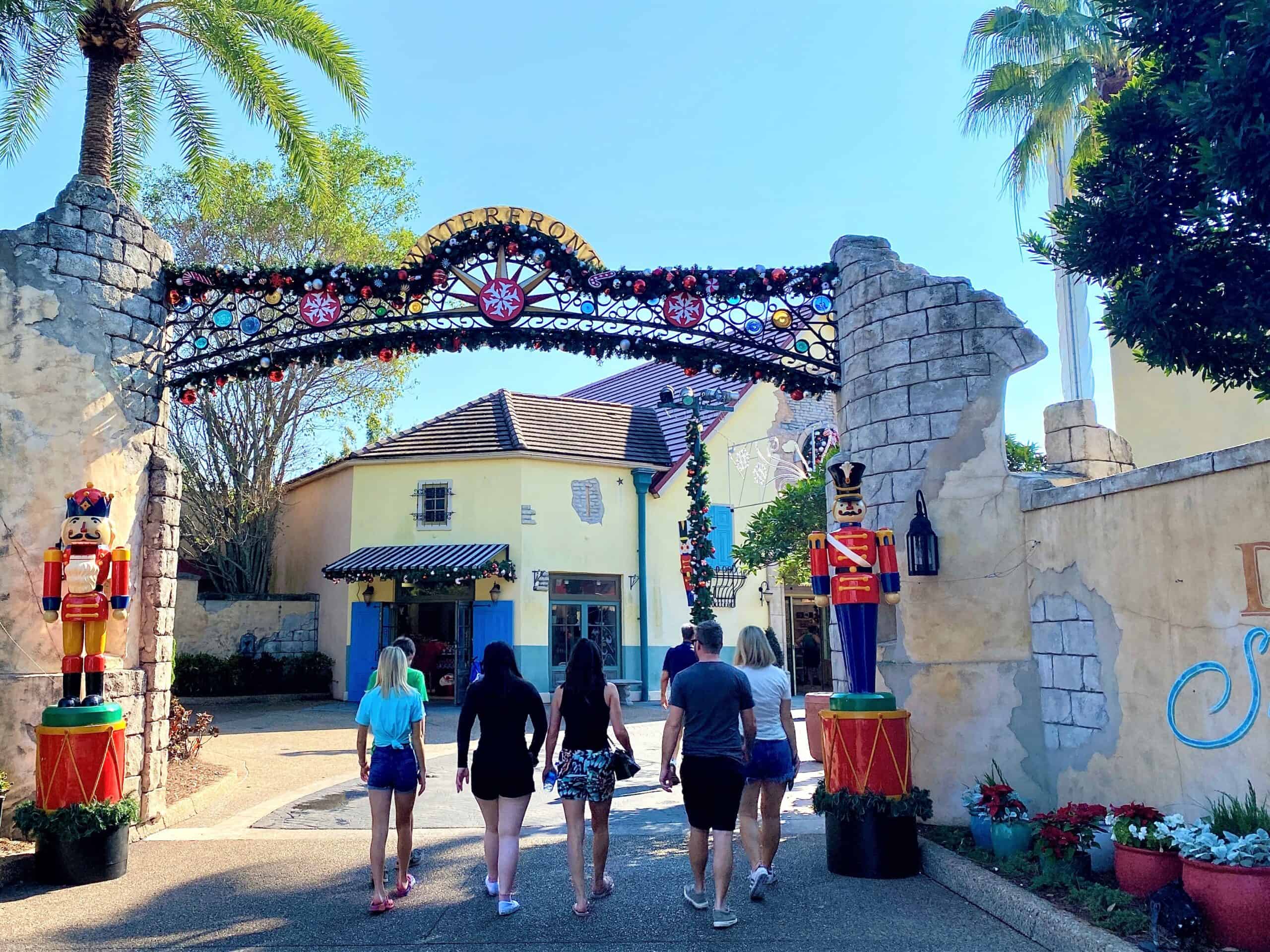 The Waterfront at SeaWorld Orlando is especially festive as a group of five casually dressed adults with their back towards the camera walk through a decorated archway flanked by two large nutcracker decorations. 