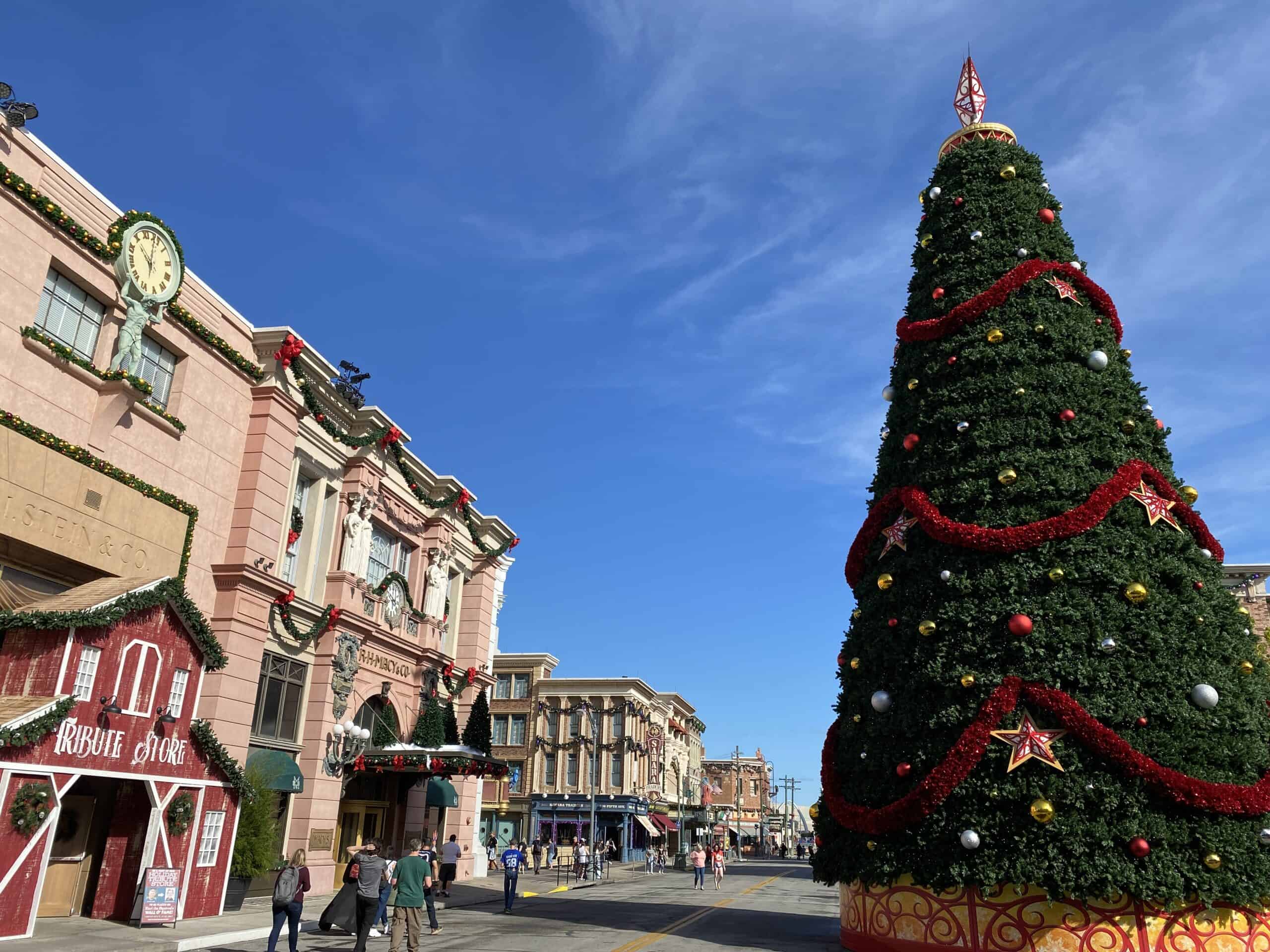 Towering Christmas Tree in New York Area Universal Studios. The large Christmas tree has red garland and red and colored ornaments, and stands tall in the right side of the photo. Large buildings and the Holiday Tribute Store are in various red colors and stand to the left of the photo. The bright blue sky is above the tree and the buildings, with just a few thin clouds