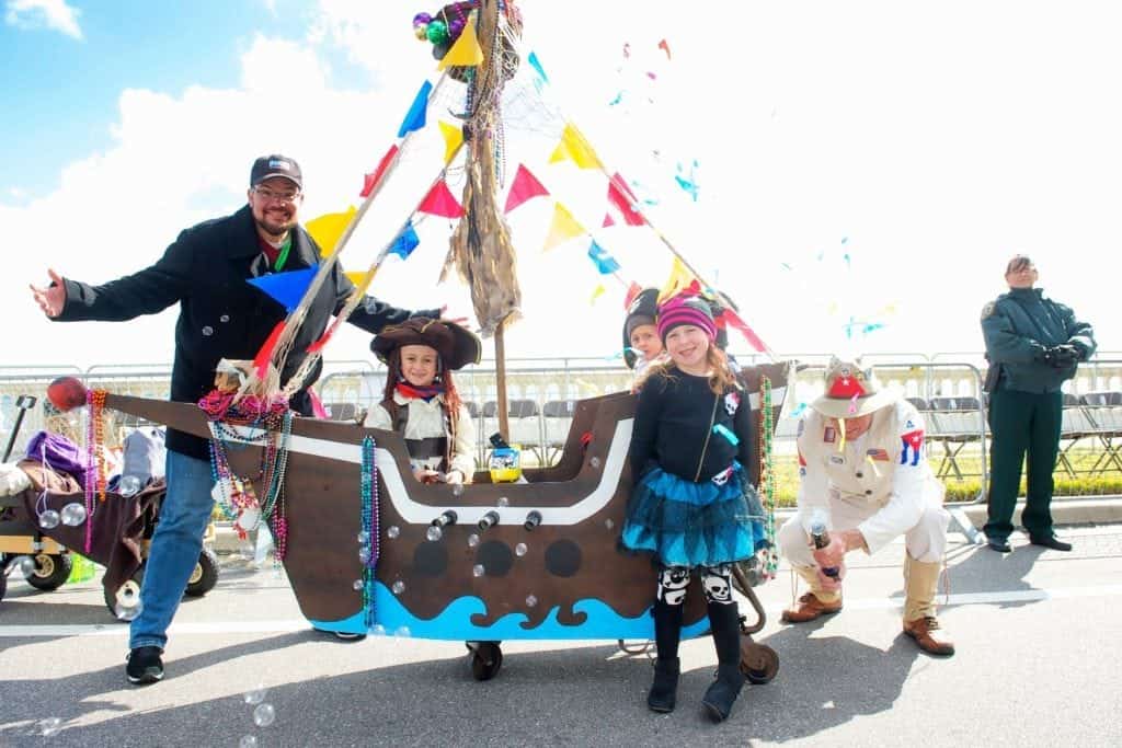 A group of parents and children dressed in pirate costumes pose with a decorated pirate ship-themed stroller at the Gasparilla Preschool Stroll, surrounded by colorful flags and festive accessories.