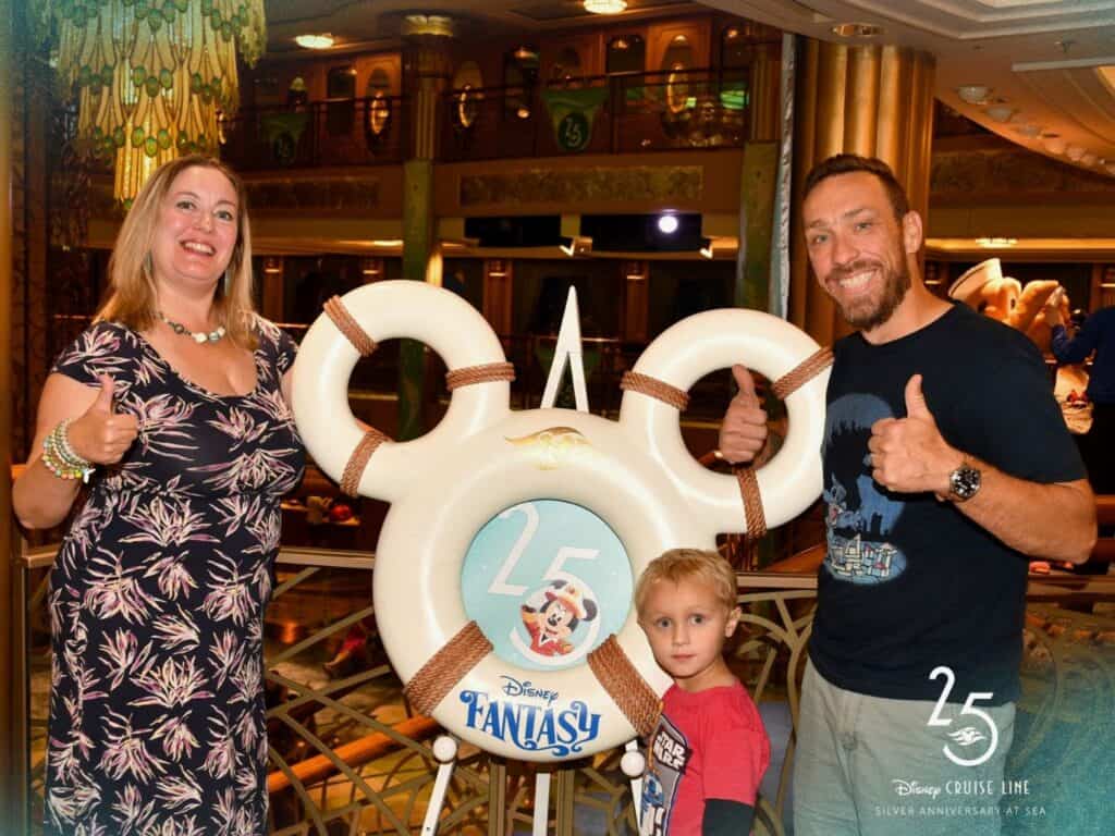 a family of two adults and one child pose in front of 25th Anniversary Disney Fantasy Cruise Photo Opportunity 