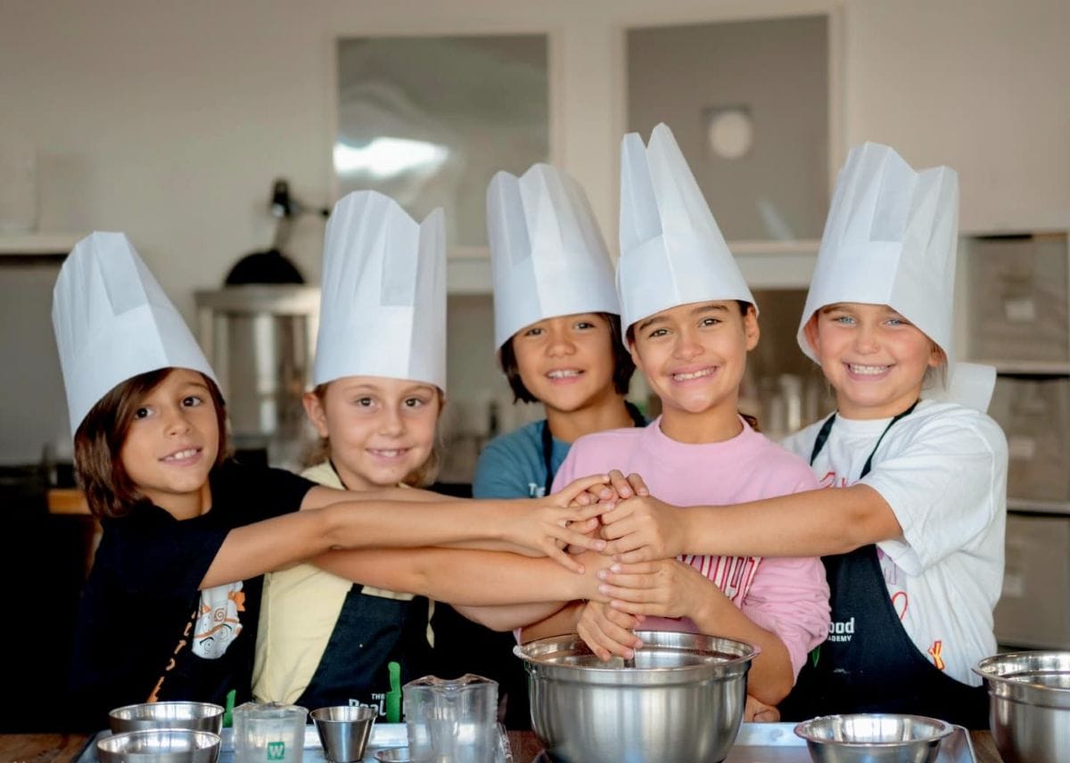 Kids wearing chefs hats smiling as they cook together at a Kids Cook Birthday party from The Real Food Academy in Tampa