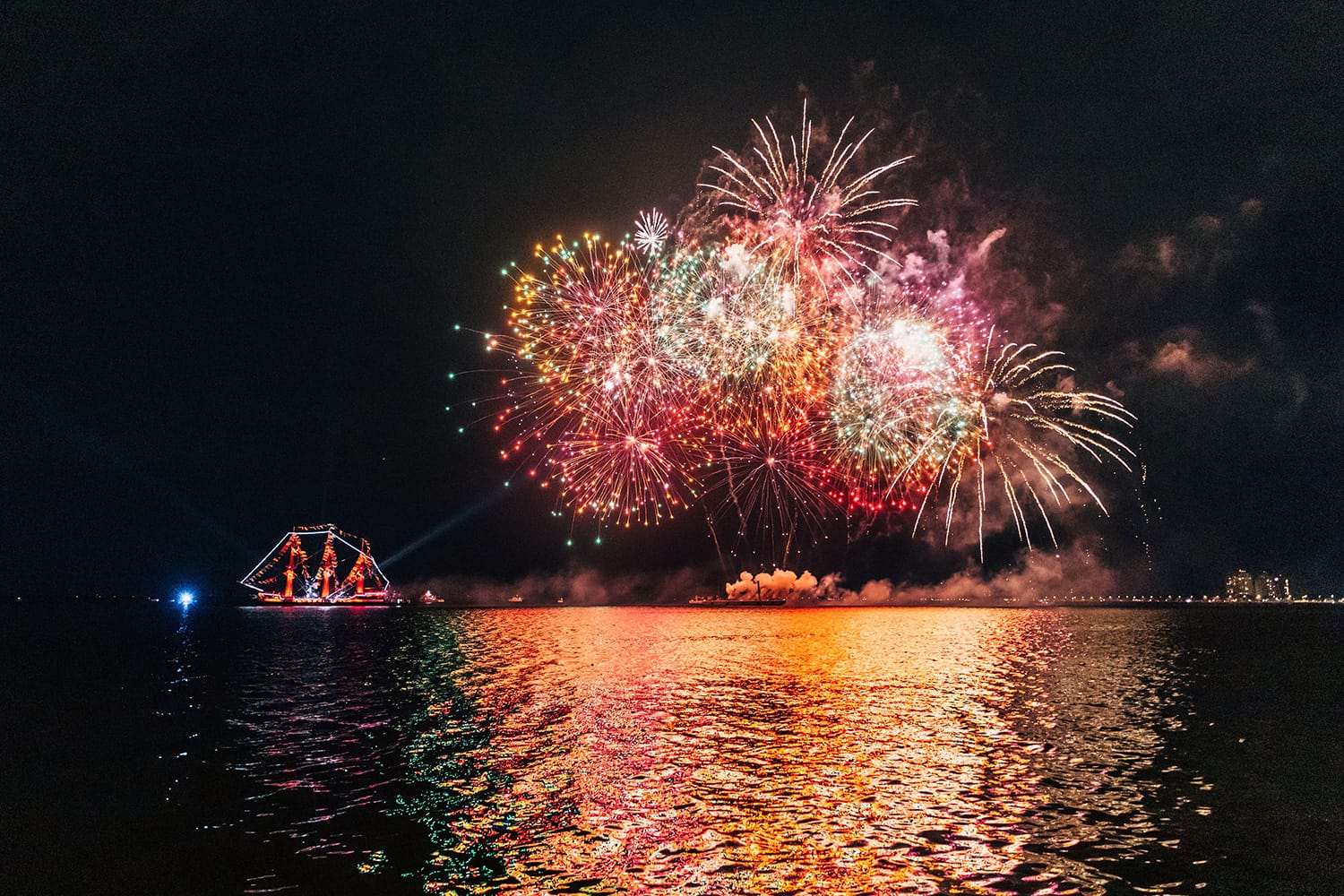 Colorful fireworks burst over the water during the Piratechnic Fireworks Extravaganza at Children’s Gasparilla, with a brightly lit pirate ship in the background.